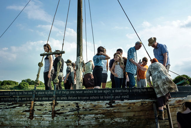 A group of people climb up and stand on an old fishing boat that is lodged in a salt marsh, and is inscribed with memories of lost things